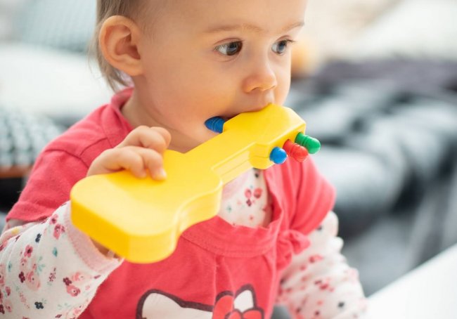 Cute caucasian 1 year old baby girl bites yellow guitar toy, concept of teething, Child bites toys